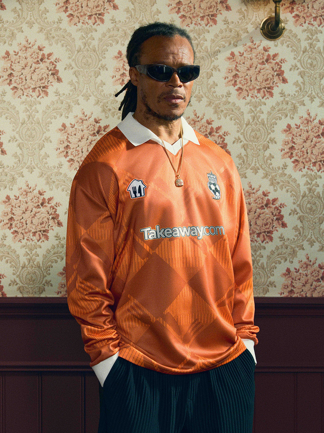 2024/25 Takeaway.com Cult Kits football jersey modeled in orange color with sunglasses against vintage wallpaper, part of Cult Kits collection.